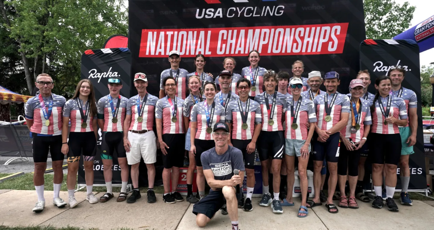 A group of riders wearing Stars and Stripes jersey stand in front of a USA Cycling National Championships backdrop.